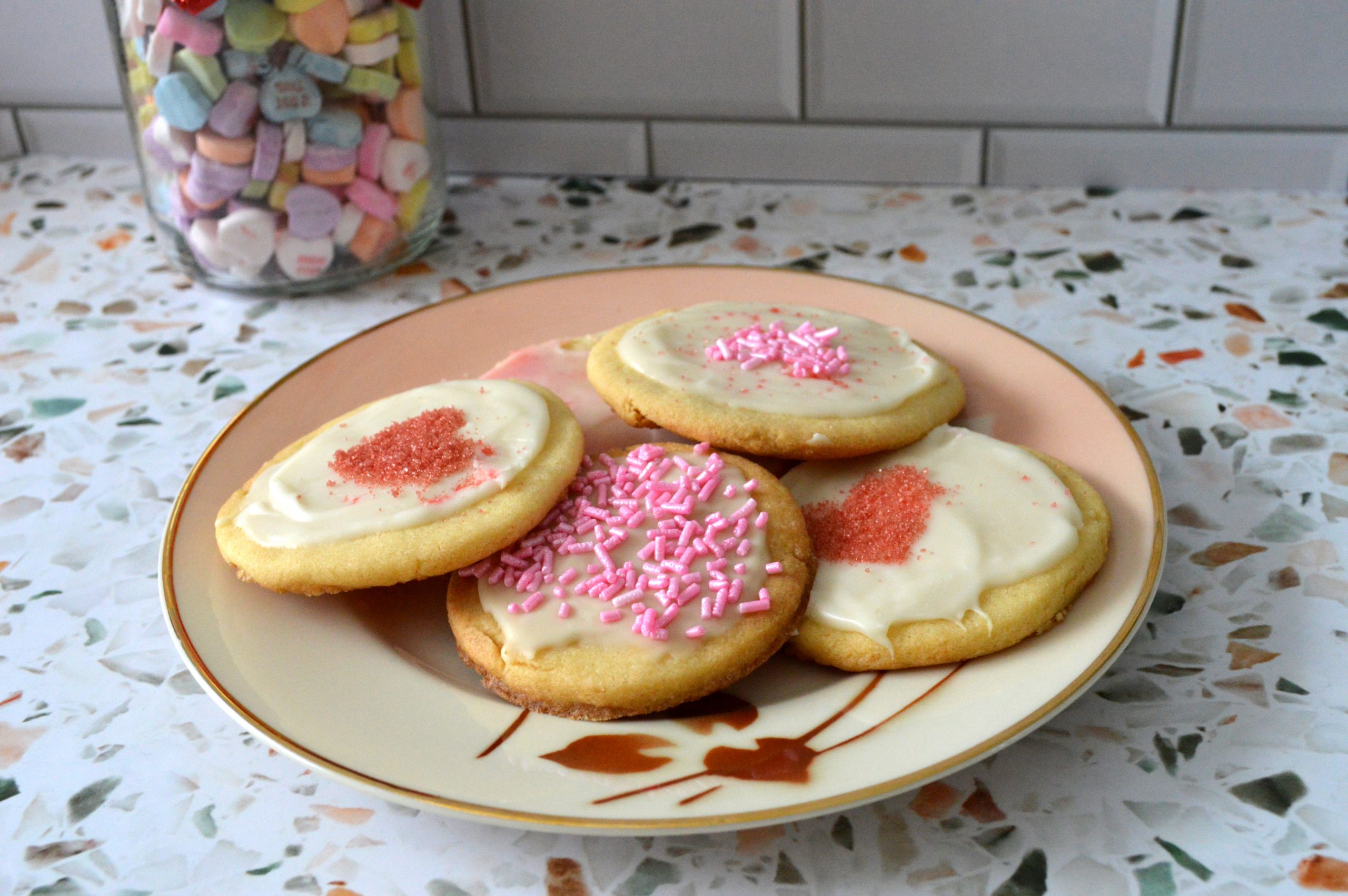 A plate of sugar cookies with white icing, pink sprinkles, and red sugar on a kitchen counter.