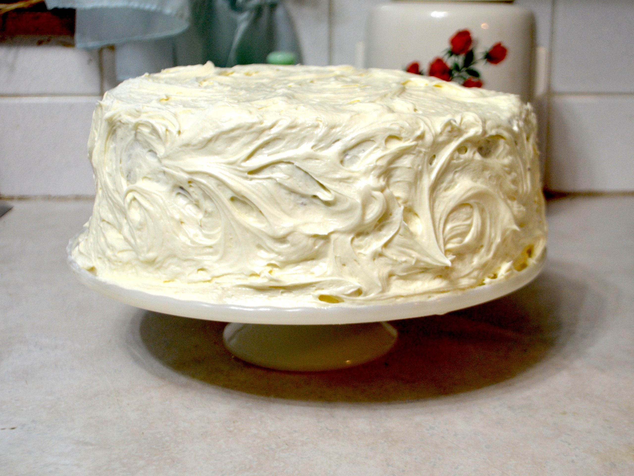 A homemade cake on a milk glass cake plate frosted with vanilla buttercream frosting.