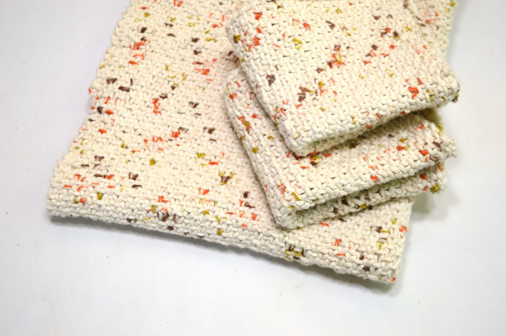 A knitted dishtowel and two knitted dishcloths in off-white yarn with orange, brown, and yellow flecks on a white background