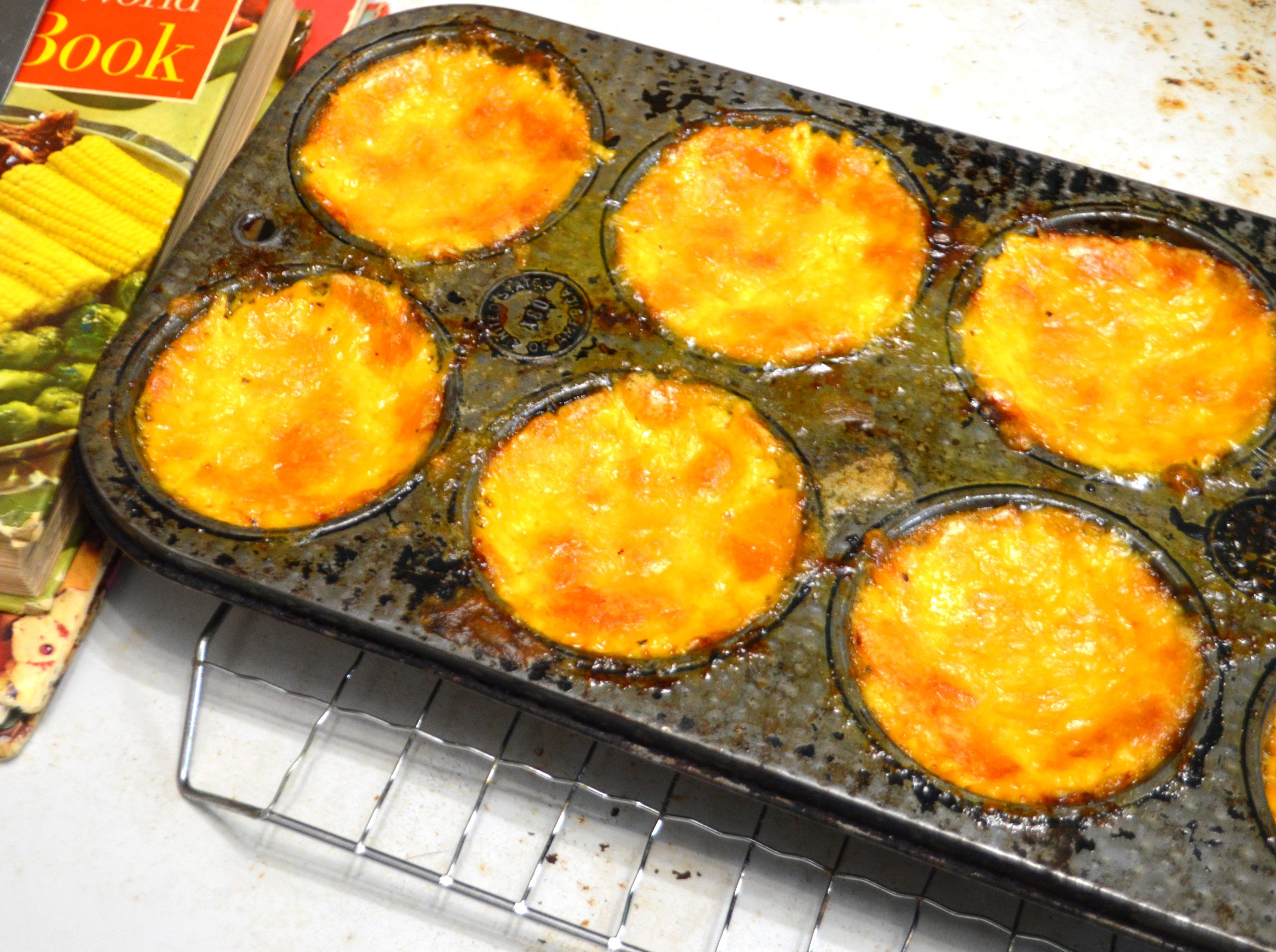 A muffin pan filled with baked cheese sits on a wire cooling rack. Vintage cookbooks are in the background.